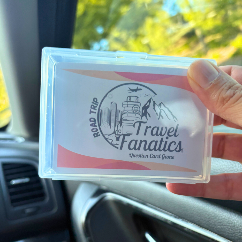 Hand holding a card with 'Travel Fanatics' road trip question card game in front of a scenic road with mountains.