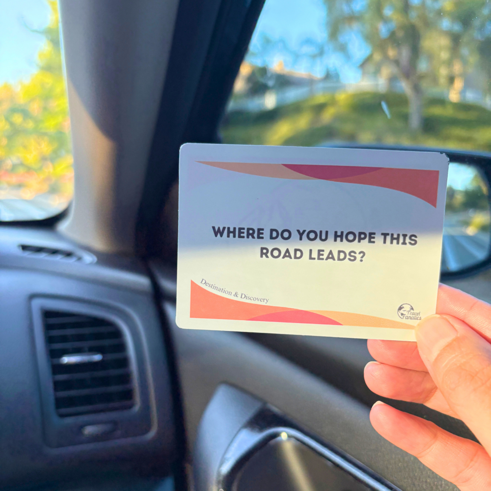 Hand holding a playing card with text 'Where do you hope this road leads?' inside a car for road trip conversations and games