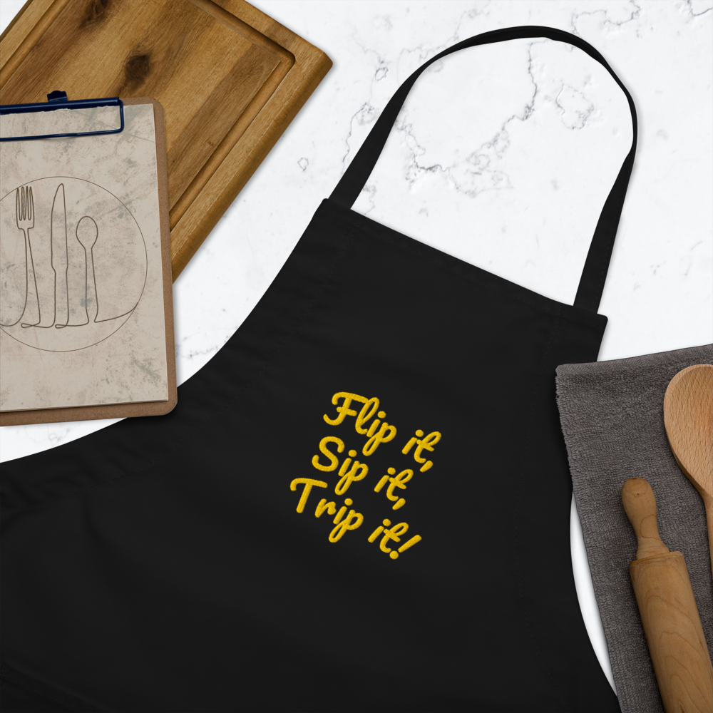 Black apron with yellow text 'Flip it, Sip it, Trip it!' on a marble surface with kitchen items.