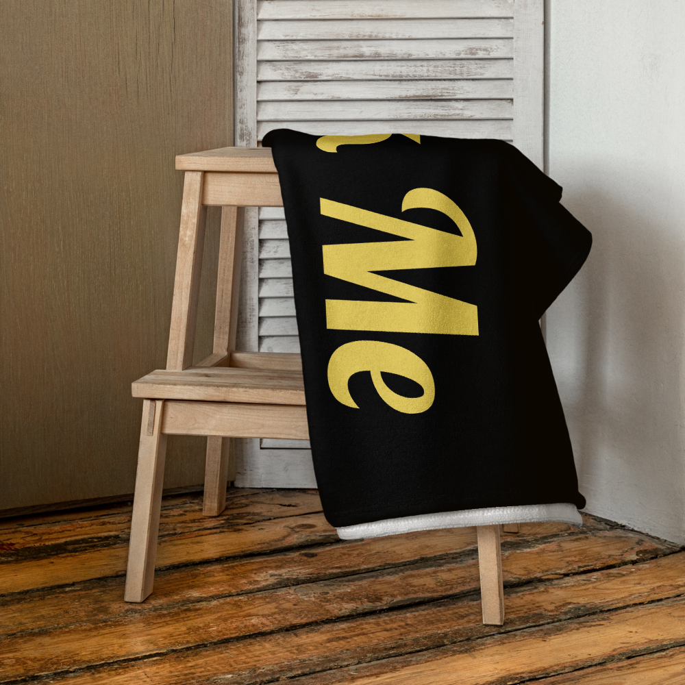 Black blanket with yellow text 'Me' draped over a wooden chair against a neutral wall.