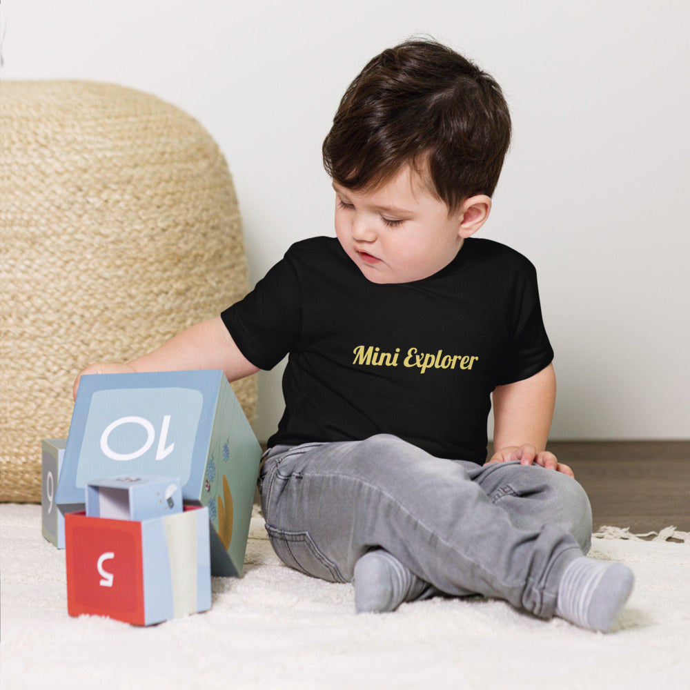 Child wearing a black t-shirt with 'Mini Explorer' text, sitting on a white surface with educational toys.