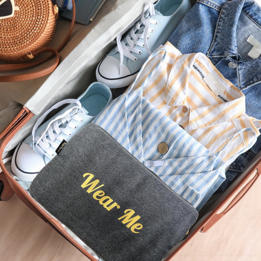 Open suitcase with clothes and a pouch labeled 'Wear Me' on a wooden floor.


