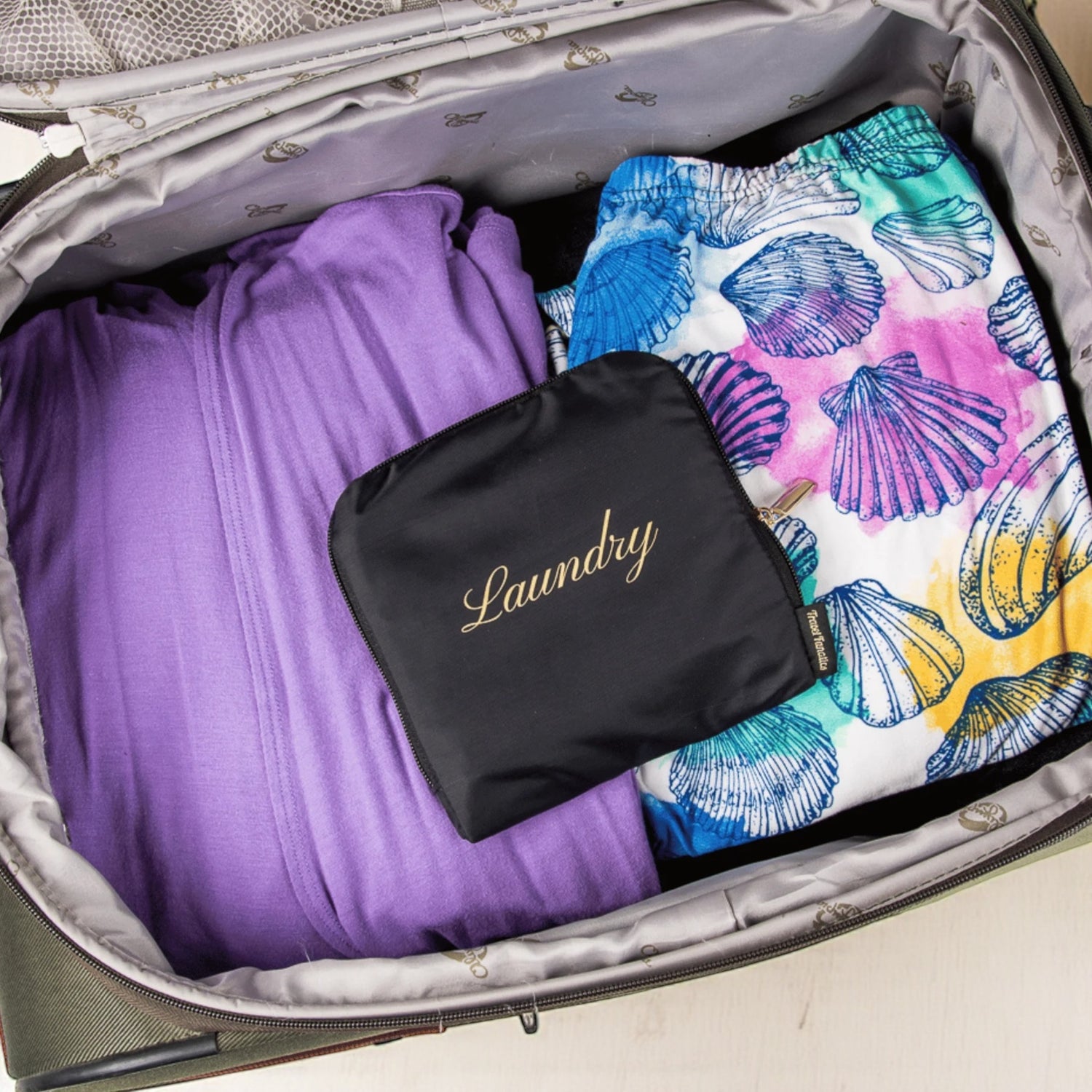 Open suitcase with purple garment, black 'Laundry' bag, and colorful shell-patterned towel.

