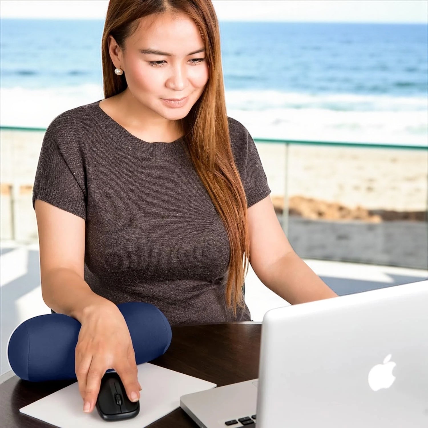 Woman using a computer with a blue pillow on her lap by the beach

