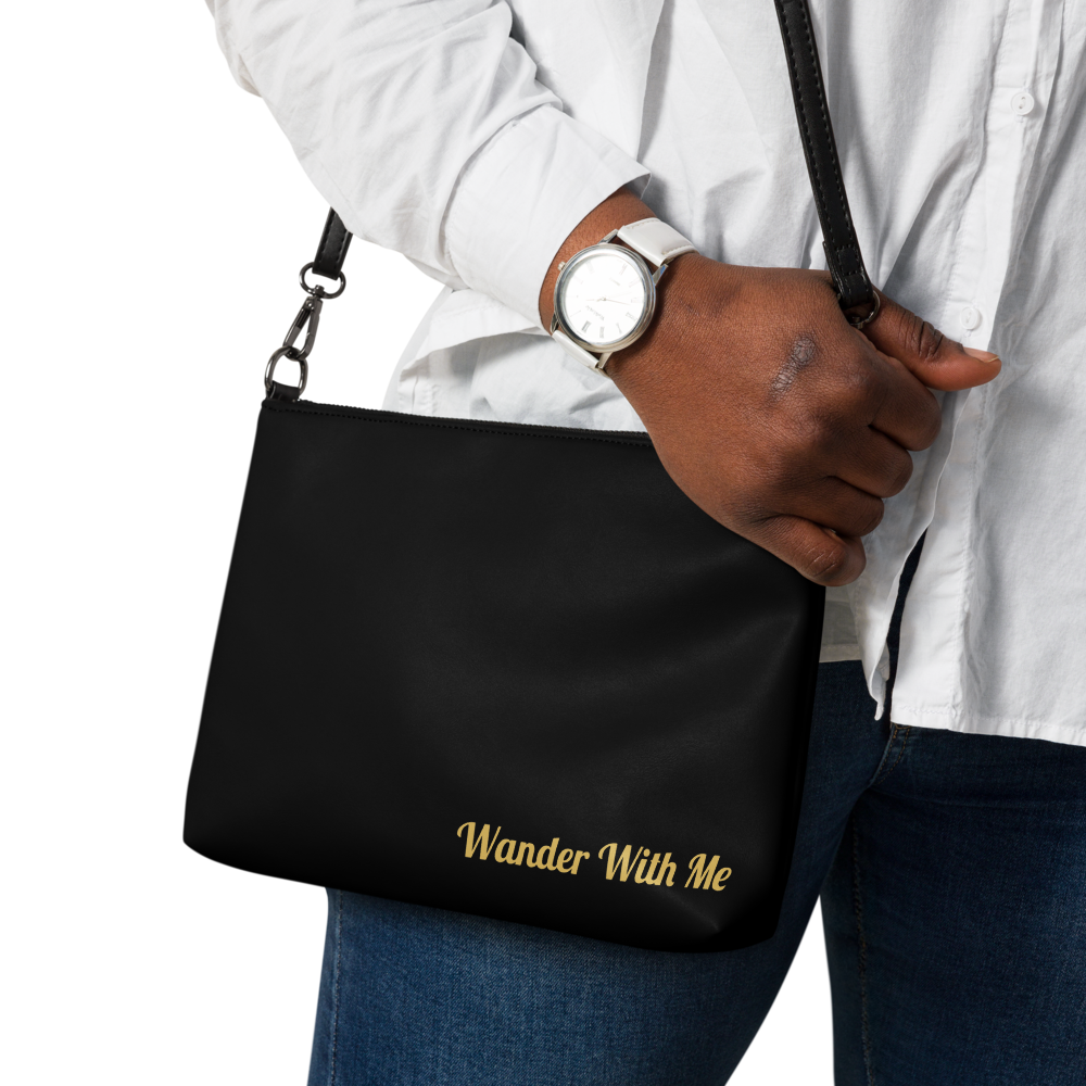 Person holding a black bag with 'Wander With Me' text, wearing a white shirt and blue jeans.

