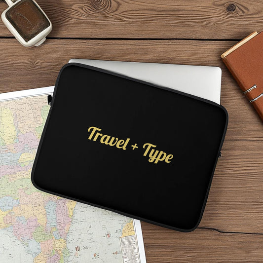 Black laptop sleeve with 'Travel + Type' text on a wooden surface with a map and notebook.

