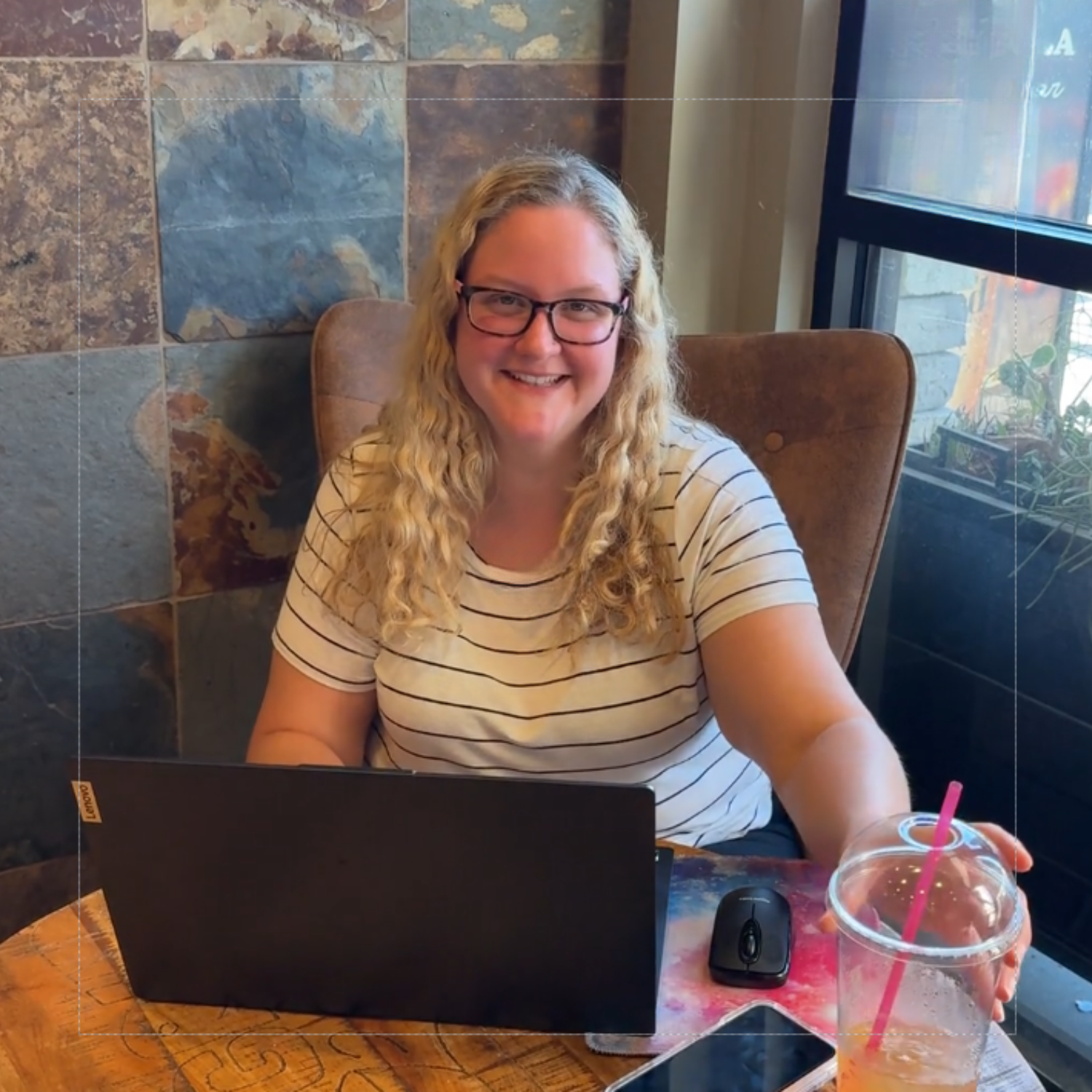 Woman sitting at a table with a laptop, phone, and drink, smiling.

